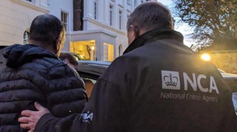 Anonymous officer of the National Crime Agency walking beside a suspected people smuggler, who is also anon, with his hand on his back. The suspected people smuggler is wearing a black puffer jacket. The NCA officer is wearing a branded black coat. 