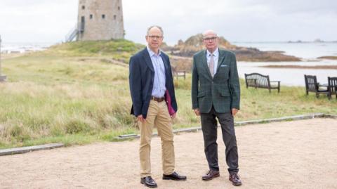 Chris Ambler and Marcus Troy are standing next to each other. They are smartly dressed. A grass area is in the background with benches overlooking the water.