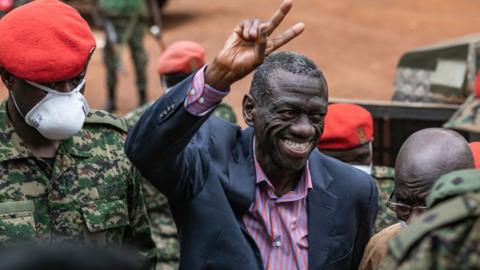 Kizza Besigye in a stripped purple and orange shirt and suit jacket smiles and pulls the victory sign as he arrives a military court surrounded by soldiers, some wearing red berets - Kampala, Uganda - 20 November 2020