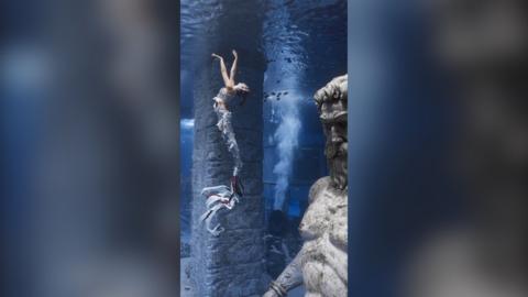 A woman in a mermaid costume reaches her arms upwards underwater, with a male statue in the foreground.
