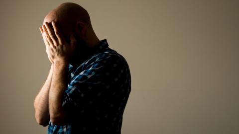 A man wearing a blue-checked shirt with his face buried in his hands