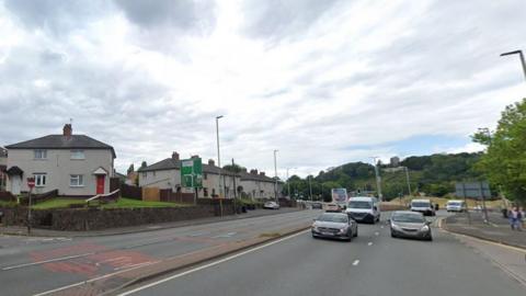 A dual carriageway with a junction with a smaller road on the left. There is a roundabout and a tree-covered hill in the background.