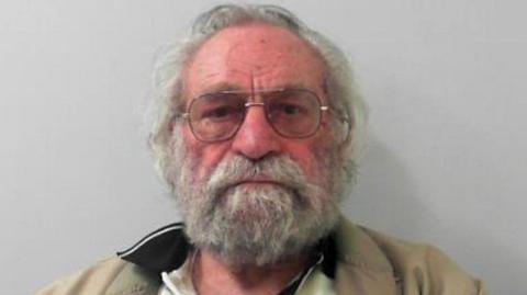 Christopher Pattinson is facing the camera. He has grey hair and a grey beard and is wearing glasses. He has a white shirt with a black collar and is also wearing a brown jacket