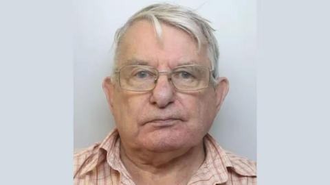 A police mugshot of an elderly man with grey hair and glasses