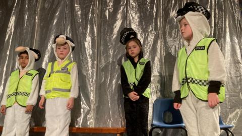 Four children are on stage. They are dressed in sheep costumes but also with police hi-vis vests and police hats.