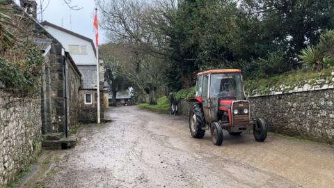 A very muddy road in Sark, with an old-fashioned red tractor on the right-hand side. There are stone walls on either side of the road with shrubbery and trees behind them. There is a building with a flag in front of it to the left.