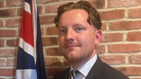 Reform UK's Chris Eynon stands in front of a brick wall and Union Jack flag wearing a black suit, striped tie and blue checked shirt.
