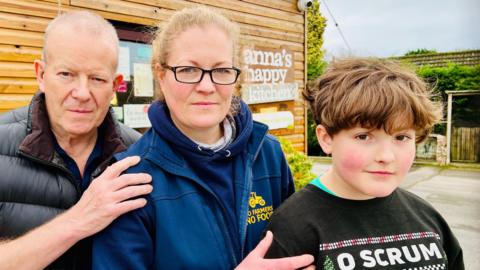 Three generations of a family stand side by side with hands on one another's shoulders. To the left is a 71-year-old grandfather, who has cropped hair and wears a black jacket; in the centre is a 42-year-old mother who has ginger hair, tied back, and wears a blue fleece with a yellow logo that reads "no farming, no food"; and to the right is an 11-year-old boy who has brown hair and wears a black Christmas jumper. They are standing in front of a wooden building with a sign reading "Anna's happy kitchen".