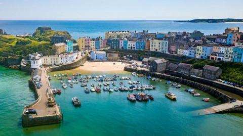 Around 80 small boats moored in green-blue water in the harbour in Tenby, viewed from above in bright sunshine. There are rows of multi-coloured houses behind the harbour - green, blue, yellow, turquoise, orange and pink - and a view out to sea at the top of the frame.