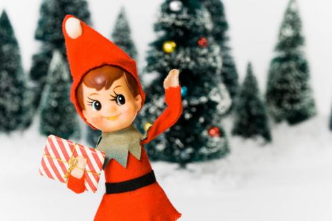 An elf in a red suit is waving with snow and trees in the background.