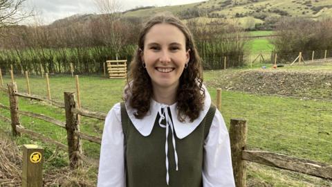 A young woman standing near a fence by a field smiling at the camera. She is wearing a white blouse and green jumper and has brown curly hair. 