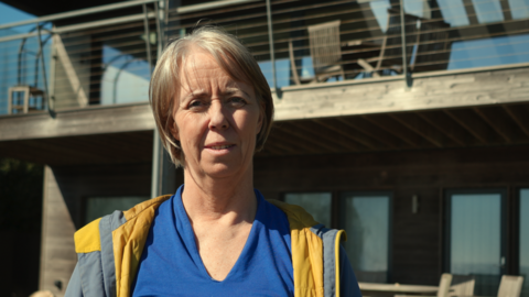 A head-and-shoulders picture of Sophie Marple, who is wearing a grey and yellow jacket over a blue top. She is standing in front of a house, although only a small portion of the house is in the image. She is looking directly into the camera.