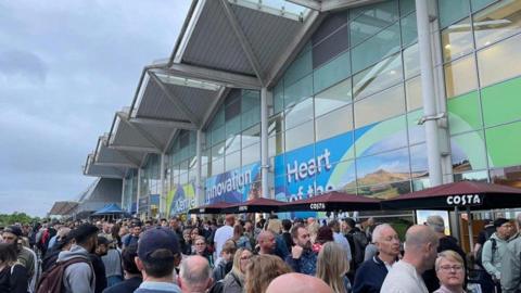 Many people are at the bottom of the photo outside an airport terminal building. Across the image is the building, with a wavy roof  and printed vinyl graphics across the front. 