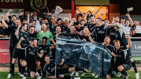 A group of men in black football kit look at the camera and celebrate a win. Some have their hands up in the air. Some are also holding up an image of a Mercedes Formula 1 driver. 