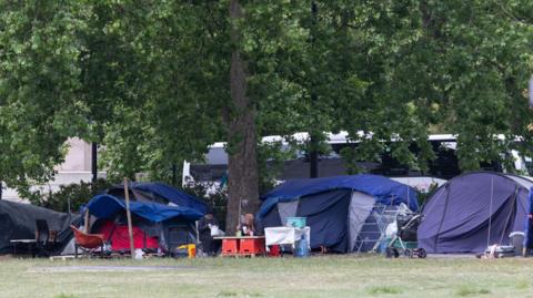 Tents and makeshift shelters are set up under large leafy trees in a grassy area, with various items such as chairs, tables, and a stroller visible. Two people sit at a small table near the centre of the image. A white coach bus is partially visible in the background behind the trees.