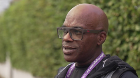 Wayne George in a black t-shirt and jacket. He wears a purple lanyard and has brown framed glasses.