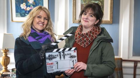 Two women stand holding a film slate. One is wearing a purple scarf and dark sweater while the other has a red checkered scarf, red shirt and green overcoat