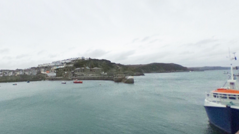 The Cornish Fishing port of Mevagissey. White houses dot a hillside, while a granite pier juts out into the sea, with cliffs behind. A blue and white boat is moored in the foreground.
