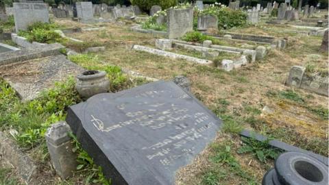 Headstones and graves in a graveyard. The headstone in the foreground has fallen over and is embossed with writing on dark coloured stone.