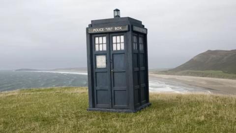 The TARDIS stands on a grassy hill overlooking a beach.