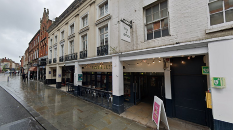 The outside of a white building in Bedford High Street. Outside the building is a hanging sign for the Slug & Lettuce. There is black writing on the front of the building which reads SLUG & LETTUCE and there are fairy lights hung up. In the same building is a nightclub where a sign hangs for it, VOGUE.