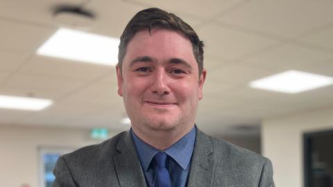 Mayor Chris Cooke has short brown hair and a grey suit and blue shirt. He has a blue tie. He is clean-shaven and standing in a classroom which is blurred out.