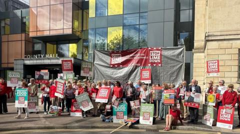 A crowd of people in red T-shirts hold red signs reading "Stop Lime Down" and green signs reading "protect food" outside a large municipal building with the words "county hall" above the door. 