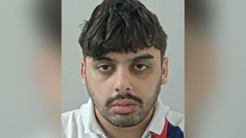 Police custody image of Jake McMahon-Bates with black hair and stubble and moustache wearing a white polo top.