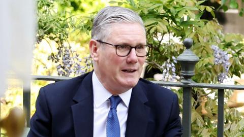 Keir Starmer smiles slightly while looking away from camera with green foliage behind him, pictured outside at Downing Street on 9 May