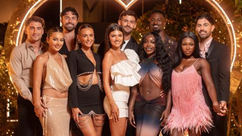 Ten Love Island contestants stand in front of a neon lit heart shape outside the Love Island villa. The photo is cropped so the ten people fill the image and they're standing as five couples, with the five men in the back row and the five women in front of them. They're all dressed smartly, with the men most in shirts and jackets and the women all in dresses.