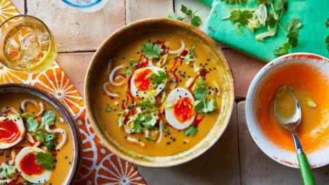 Three bowls - two with soup and toppings including a boiled egg, one empty with a spoon in it - sit on a brick surface