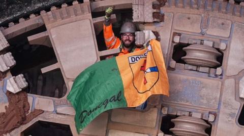 A huge metal circle is protruding out of a piece of land. The metal is bronze in colour. A man in an orange high-vis jacket and grey helmet is poking his head out with his thumb up in the air. He is holding a Donegal flag, which is green and orange.