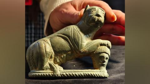 A close-up of a Romano-British vehicle fitting. It is in the shape of a big cat, resting on its hind legs with its paws resting on a severed bearded head. It is being held up by a woman's hand. Found at Epping Forest, near Harlow, Essex.