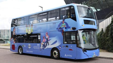 A blue double-decker Transport for London bus parked on the road, viewed from the front, side on. The design of the bus is coloured as a gradient from purple-blue at the bottom to light blue at the top. Designs symbolising the history of Transport for London are shown on the side of the bus, including the rollout of accessible buses in 2005. These events are symbolised by cartoon drawings of people, integrated with the Transport for London roundel.