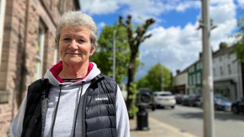 Margaret Morgan standing on Main Street in Cockermouth. She has short grey permed hair and is wearing a grey hoodie with pink lining and a black bodywarmer. She is looking at the camera with a neutral expression. The background is blurred and there is a pollarded tree behind her, cars and shops.