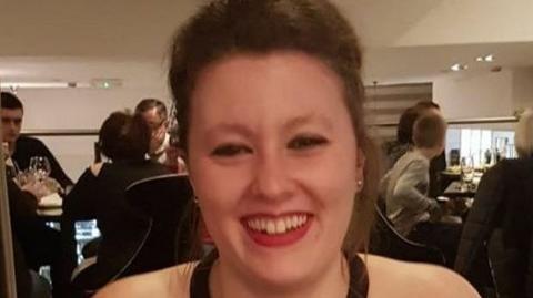 Woman with dark brown hair pinned back wearing stud earrings and a black halterneck with red lipstick smiling in a restaurant as other diners are in the background  