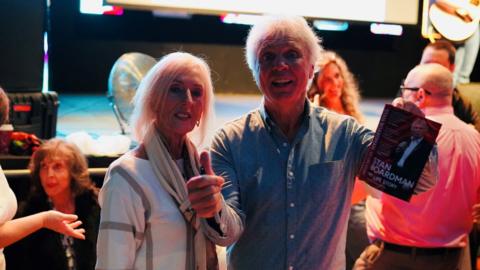 Comedian Stan Boardman stands with a woman in front of a stage. He is holding up a book which is called 'My Life Story' and doing a thumbs up with his other hand. 