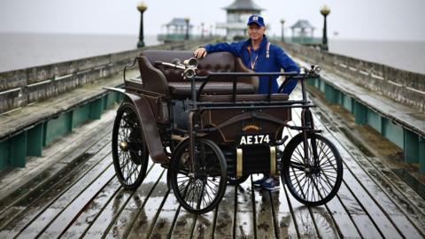 A rainy Clevedon Pier, with Mark Reber standing beside a brown wooden framed Victorian motor car