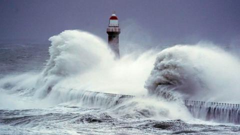 A huge wave crashes over a sea wall and lighthouse against a deep blue sky