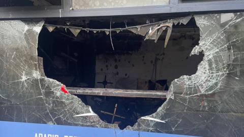 Picture shows a large broken hole in a window at the front of a cafe in Belfast - the picture focus on the large hole in the middle of the window, which shows most of the window gone. At the edges, are broken sharp crags of the remaining glass in the frame. Inside the cafe has been destroyed by fire.