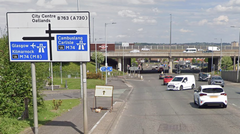 Road junction with overhead bridge and multiple cars, featuring directional road signs pointing to Glasgow, Kilmarnock, Cambuslang, Carlisle and nearby city centre routes.