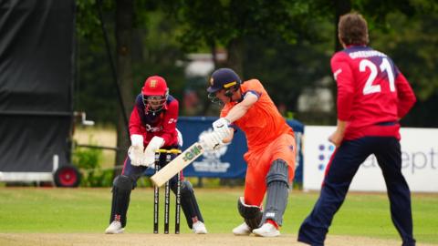 A Netherlands batter, wearing orange, attempts to hit the ball as two Jersey players, wearing red and navy blue outfits, watch on