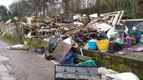 A large pile of waste including bits of wood, plastic tubs and other unidentifiable waste. Trees in the background and a lane in the foreground.