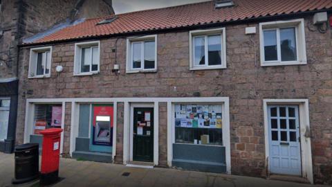Wooler Post Office. It is a stone building with two storeys. A cashpoint is next to the door and a post box is positioned on the pavement.