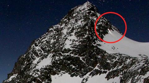 Mountain against a night sky with a red circle highlighting torchlight on the side of the mountain.