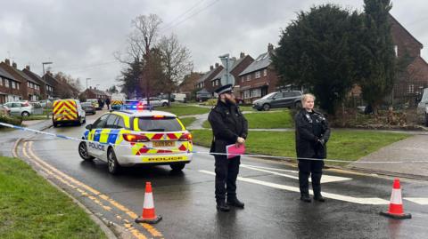 Two police officers stood behind the cordon closing off Hazel Avenue, Guildford, between the junctions with Lime Grove and Willow Way. Several police cars and a van can be seen in the background further up the street.