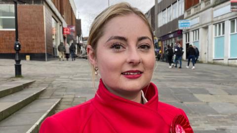 A woman in a red coat with a red rosette on a lapel. She has blonde hair and is standing on a pedestrianised shopping street. 