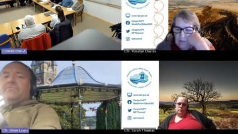 A screen grab of the broadcast of a council meeting, with four windows divided by a blue council logo on a white background. The top left window shows the council room with people at a table, another on the right shows a woman with glasses and her hand on her cheek with a dark background and below that a woman with glasses and a screensaver in the background. Councillor Lewis is in the bottom left looking down at the camera with a scenic pavilion background photo. You can just see he has a seatbelt across his chest 