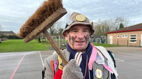 Michael Czarnobaj is wearing a tweed hat and tweed jacket with badges on. The jacket has extra ribbons pinned to it and he has a red, white and blue scarf around his neck. He has some black "soot" smeared on his cheeks and is holding a broom with the head near his. He is standing on a school playground.