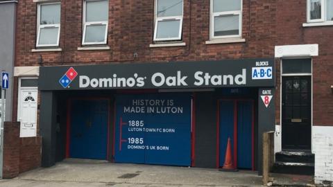 Photo of Luton Town FC's Oak stand entrance. It is branded with Domino's Pizza logos and there are terraced houses on either side of the entrance